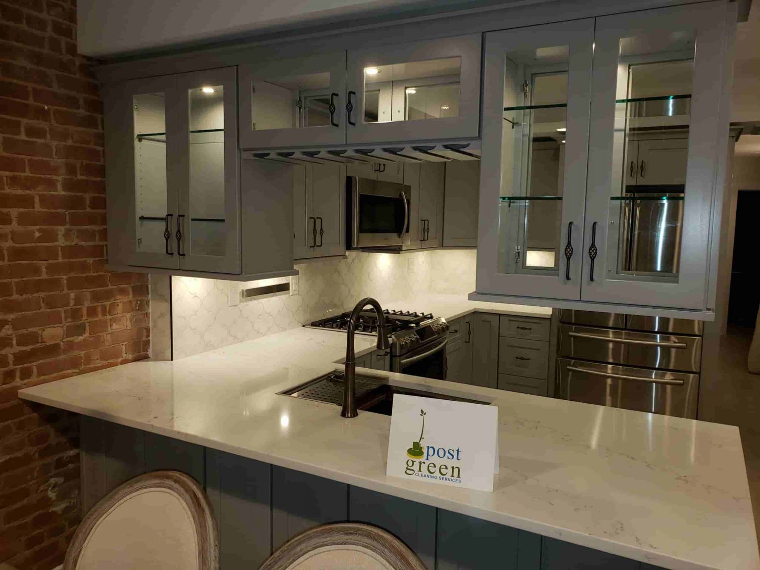 A grey-cabinet kitchen with exposed brick wall, white quartz counter, and stainless appliances